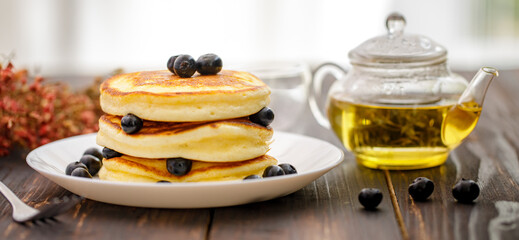 Sweet food. Stack of delicious pancakes with fork rests blueberries and honey, teapot in white plate on blur wooden background.