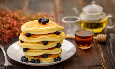 Sweet food. Stack of delicious pancakes with fork rests blueberries and honey, teapot in white plate on blur wooden background.
