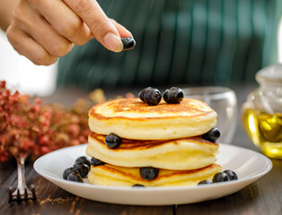 Close up hand of unrecognizable woman holding hand decorating Pancake by picking up blueberries and blur teapot background. Concept sweet food.