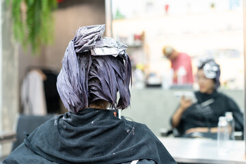 Focus of back view young Asian woman sitting in the hairdressing chair using a cell phone reading news while having hair coloring process with blurred background of herself in a mirror in a hair salon
