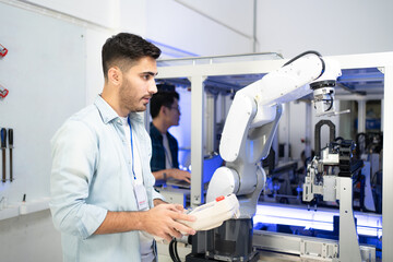 Young Caucasian male industrial engineer in a blue shirt standing controlling an automation robotic arm machine for multiple industrial applications with a robot controller in an industrial factory.