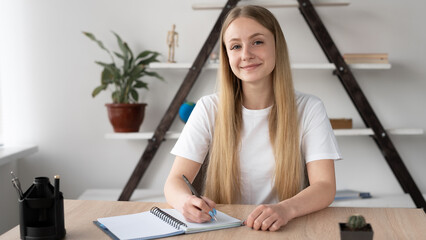 Portrait of a blonde girl with long hair sitting at a desk in the office with an open notebook,...