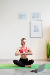 Fototapeta premium Yoga practice. Sportive woman. Online training. Harmony mind. Smiling athletic lady in sportswear in lotus pose namaste hands before laptop in light room interior.