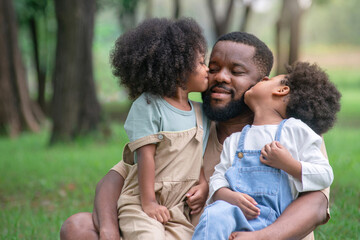 African father and daughter and son had a great time at the outdoor park, two children are sitting on the lap of the father and kiss him, Father's day