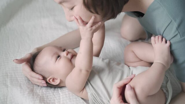 asian woman mother play and kissing baby infant on cheek softly on bed. baby happy when mother parent kissing with love and care. mom using nose touching kiss daughter at home. authentic lifestyle.