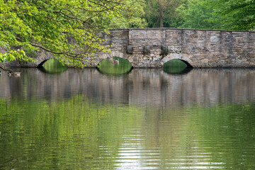 Fototapeta na wymiar Alte Steinbrücke mit zwei Bögen über einen Wassergraben. Wunderschöne grüne Spiegelungen von den Bäumen mit frischem Frühlingslaub auf dem Wasser.