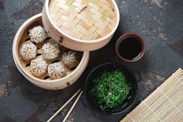 Fototapeta na wymiar Bamboo steamer with chinese soup dumplings and seaweed salad, high angle view over brown stone background, horizontal shot