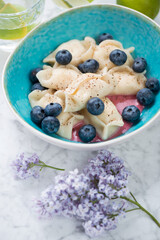 Turquoise bowl with vareniki or curd dumplings and bilberry, vertical shot on a light-grey marble...