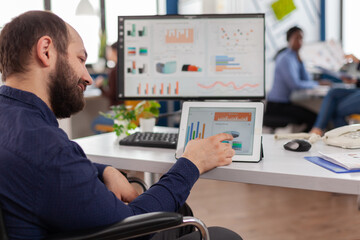 Paralysed business man sitting immobilized in wheelchair working at tablet and computer in same time analysing financial graphs, pc showing data prosessing on display for project regarding economy.