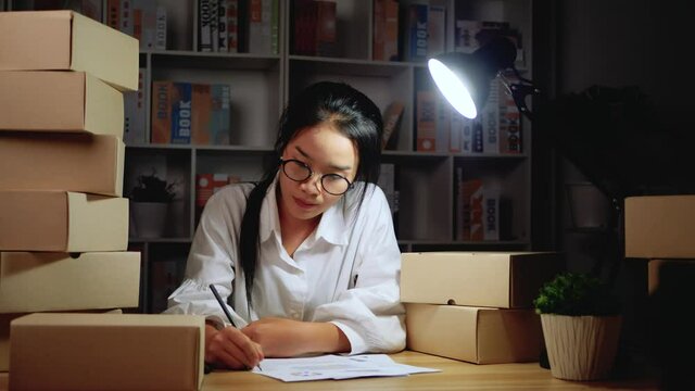 Tired startup businesswoman is packaging order in cardboard box and check list for sending in the night at home, Startup business and entrepreneurship concept. 4K