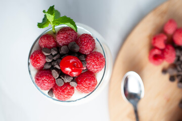 A glass of yogurt with raspberries granola, strawberries and chocolate chips with a mint or peppermint leaf on a table with wooden cutting board on the side  and a spoon  flat lay or top view.