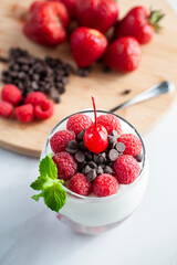 A glass of yogurt with raspberries granola, strawberries and chocolate chips with a mint or peppermint leaf and a cherry on top over a wooden cutting board in a white background vertical .