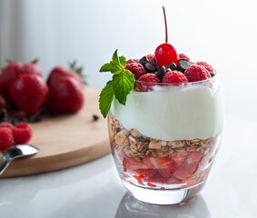 A glass of yogurt with raspberries granola, strawberries and chocolate chips with a mint or peppermint leaf on top over a wooden cutting board in a white background lit by a window