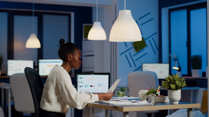 African businesswoman reading business reports from laptop in start up office late night typing new...