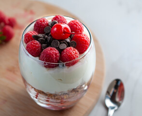A glass of yogurt with raspberries granola, strawberries and chocolate chips with a spoon on the table on the side of a  wooden cutting board in a white background, horizontal close up