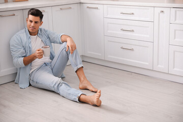 Man with cup of drink sitting on warm floor in kitchen, space for text. Heating system