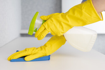 Woman in yellow rubber gloves cleaning table with blue cloth and spray.