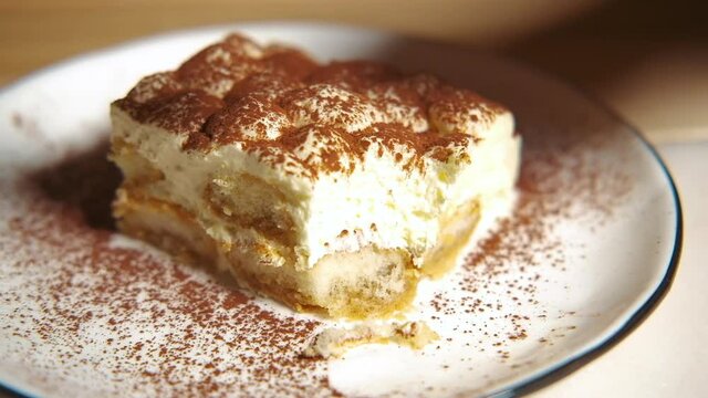 Camera slowly moves over tiramisu cake, focuses on the tiramisu cake. Slow motion. Eating tiramisu cake. Sweet traditional Italian dessert on a white plate on rustic wooden table.