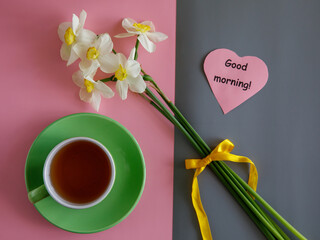 Good morning wishes note in shape heart, tea cup and white yellow daffodils bouquet on bicolor pink and grey paper trend minimalism background top view flat lay horizontal
