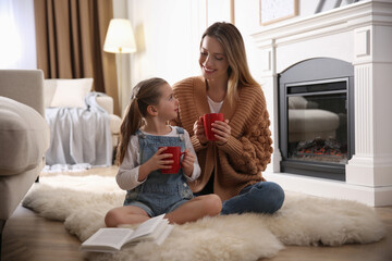 Happy woman and her daughter with cups of hot drink resting near fireplace at home