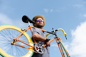 Hispanic hipster carrying a colorful fixie bicycle on his shoulder with a blue sky as background....
