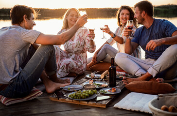 Group of friends having fun on picnic near a lake, sitting on pier eating and drinking wine.