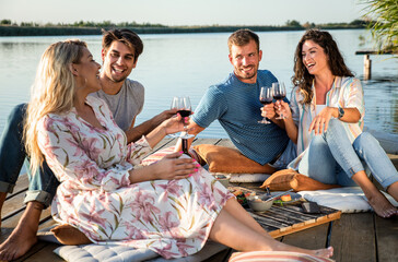 Group of friends having fun on picnic near a lake, sitting on pier eating and drinking wine.