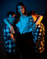 Two guys in a plaid shirt are holding a girl on a dark background in a studio with multicolored light