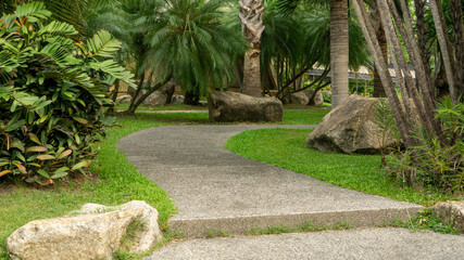 Obraz premium Walkway in garden of public park among greenery trees, flower shrub and bush, grey stone pavement walkway beside green grass lawn in a good care maintenance landscaped