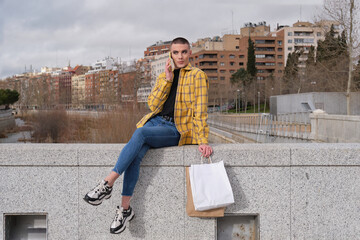 Young non binary person speaking on the smartphone with shopping bags sitting on a wall. Make up...