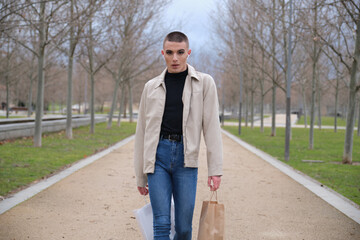 Young man wearing make up walking with shopping bags and listening music in a park. Queer person.