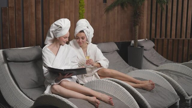 Young relaxed women in bathrobes looking at menu in spa salon, choosing procedures, having rest, communicating.