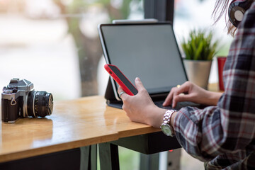 Close up of a woman hand holding a smartphone while typing on a tablet keyboard a camera a blank photo at a wooden table a coffee shop.