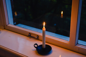 Long white candle in an old style vintage candle holder burning on the wooden big window sill in...