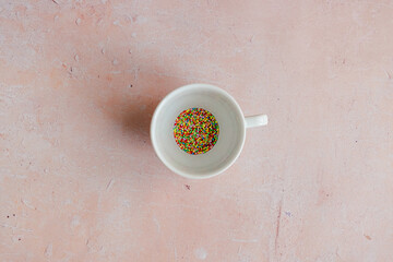 A white coffee cup filled with colorful candy sprinkles and  hundreds and thousands on a pink aged concrete counter background. Added sugar and sweetener consumption. Obesity health problem.
