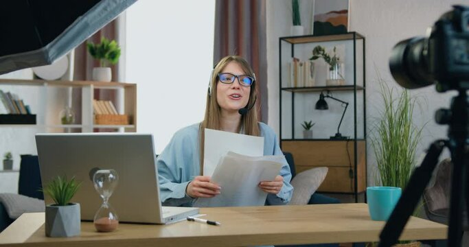 Beautiful satisfied experienced 25-aged female person in headphones throwing up financial reports during holding online webinar on camera and greeting her audience with successfully ending her course