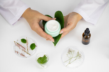 Close-up hands hold cream jar on green leaf on white laboratory background. Lab-formulated plan-based cosmetics, laboratory clinical developing skin care formulas, active natural ingredients concept