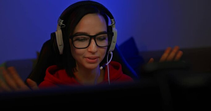 Portrait of a beautiful girl with glasses and headphones is broadcasting online over the Internet, she waves her hands and greets the participants of the online conference. Blogger sits at the monitor