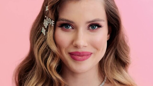 A close-up view of a smiling young woman wearing gorgeous jewelries and hairpins is looking to the camera standing isolated over a pink wall in the studio
