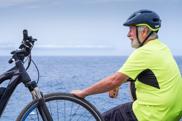 Smiling senior cyclist man resting at sea on the cliff after excursion with his electro bike.