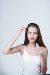 A charming girl in a white T-shirt with long hair holds a serum in her hands and applies it with a pipette to her face