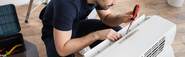 cropped view of bearded handyman holding screwdriver while fixing broken air conditioner, banner