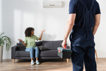 curly african american woman looking at broken air conditioner and showing shrug gesture near...