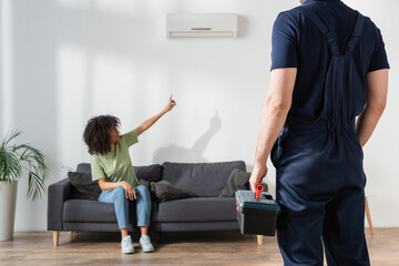 blurred african american woman pointing at broken air conditioner near handyman with toolbox