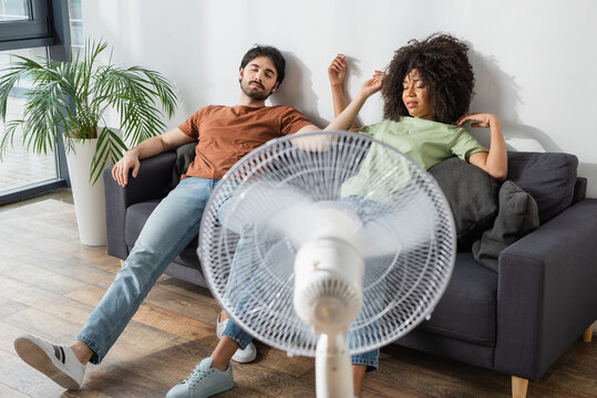 exhausted interracial couple sitting on couch near blurred electric fan in living room - Powered by Adobe