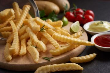 Homemade baked potato fries with mayonnaise, tomato sauce and rosemary on wooden board. tasty french fries on cutting board, in brown paper bag on black stone table background, unhealthy food.