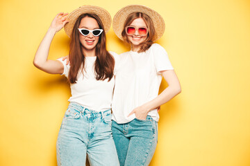 Two young beautiful smiling hipster female in trendy summer white t-shirt and jeans clothes.Sexy carefree women posing near yellow wall in studio.Positive models in hats