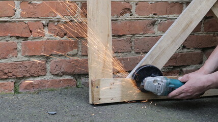 the process of work of an angle saw with flying sparks when cutting iron, male hands saw off metal parts in the frame structure with a grinder against the background of a street brick wall - obrazy, fototapety, plakaty