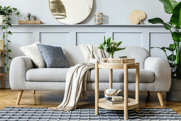 Scandinavian living room interior with design grey sofa, wooden coffee table, tropical plants, shelf, mirror, furniture, plaid pillow, teapot, book and elegant personal accessories in home decor.
