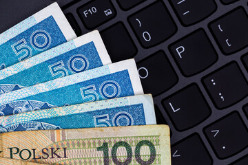 A set of Polish 50 and 100 PLN banknotes arranged on a black laptop keyboard. Photo taken under artificial, soft light